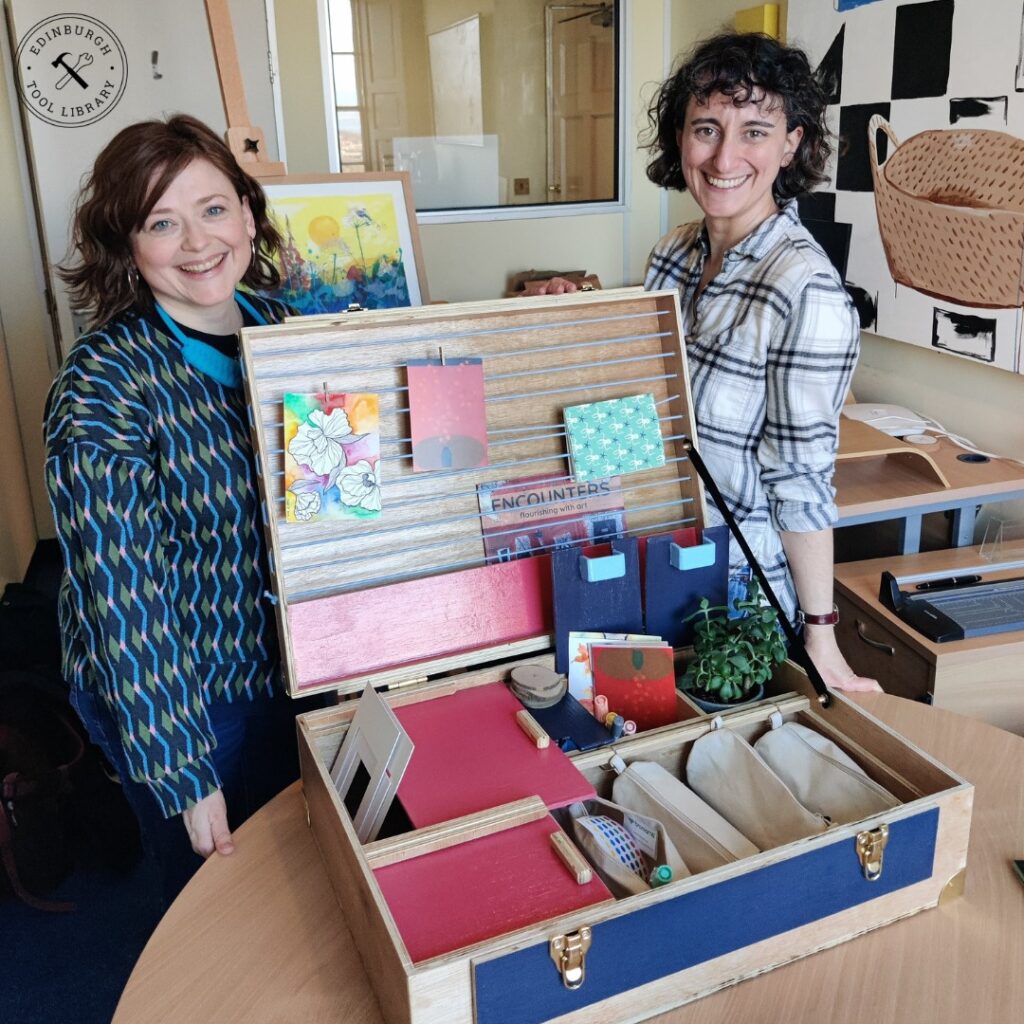 two people smiling and standing by an open wooden artbox filled with stationery supplies